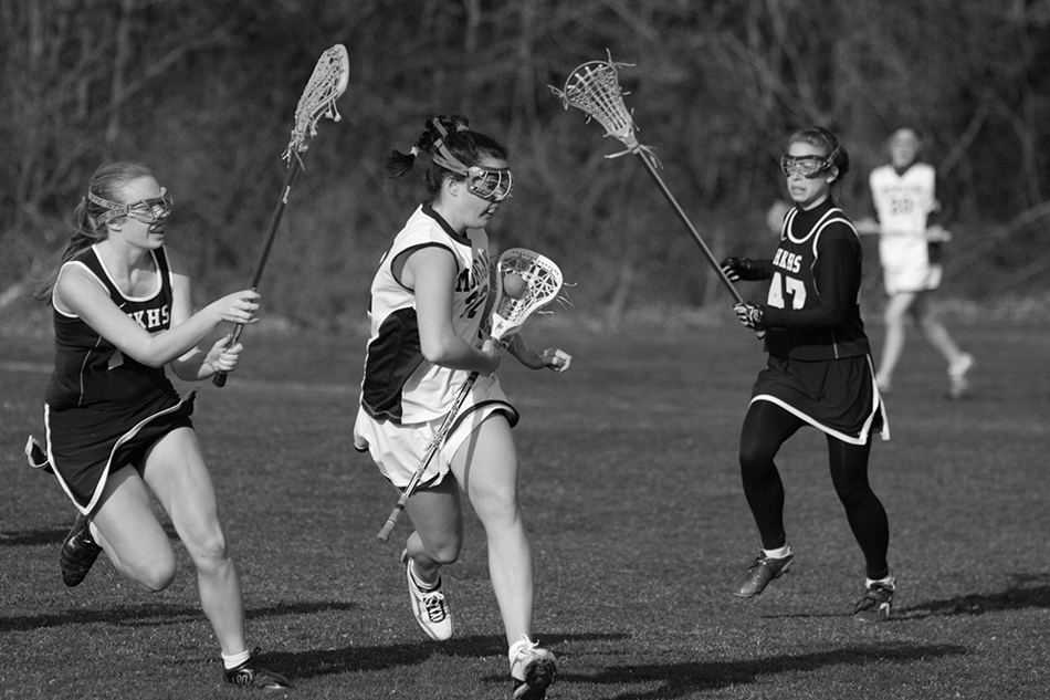 Girls’ lacrosse players in white and black uniforms are in the midst of a play.